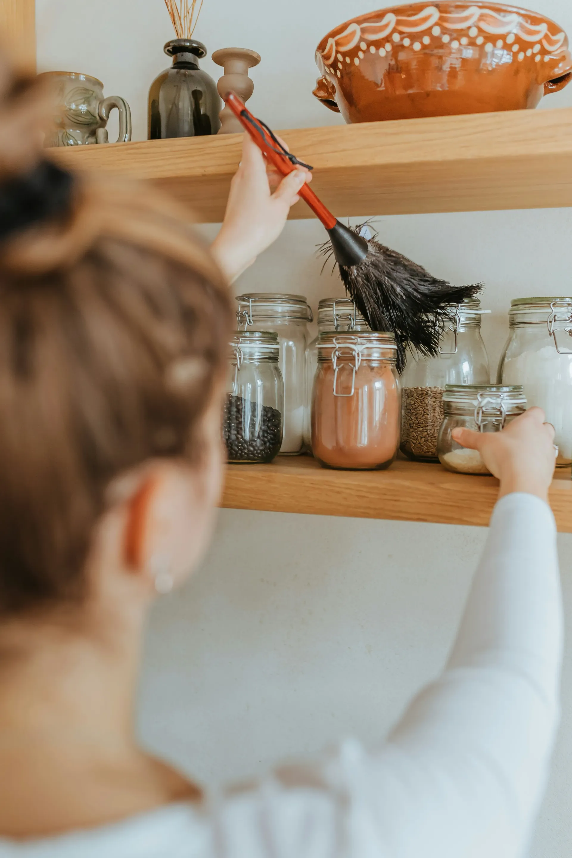 A person's arm reaching up with a feather duster to clean a wooden shelf, with several glass jars of food and decorative pottery visible.