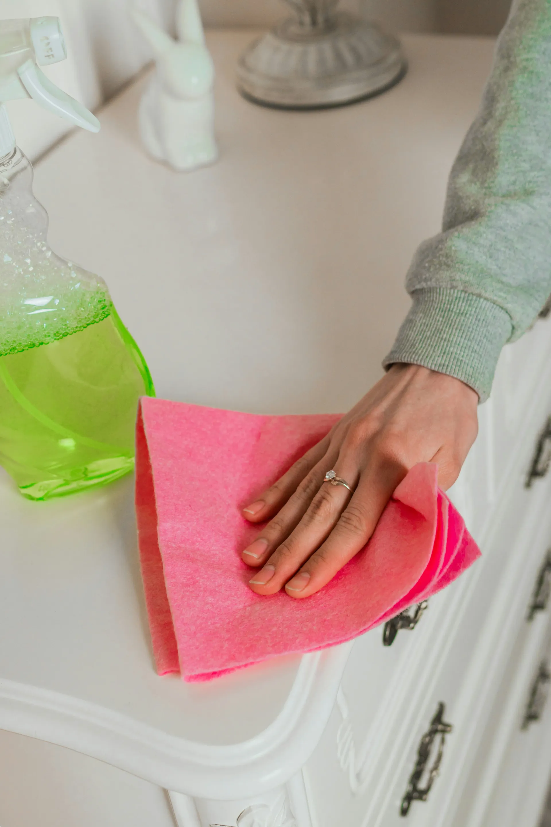 Hand cleaning a white surface with a pink cloth and green spray bottle.