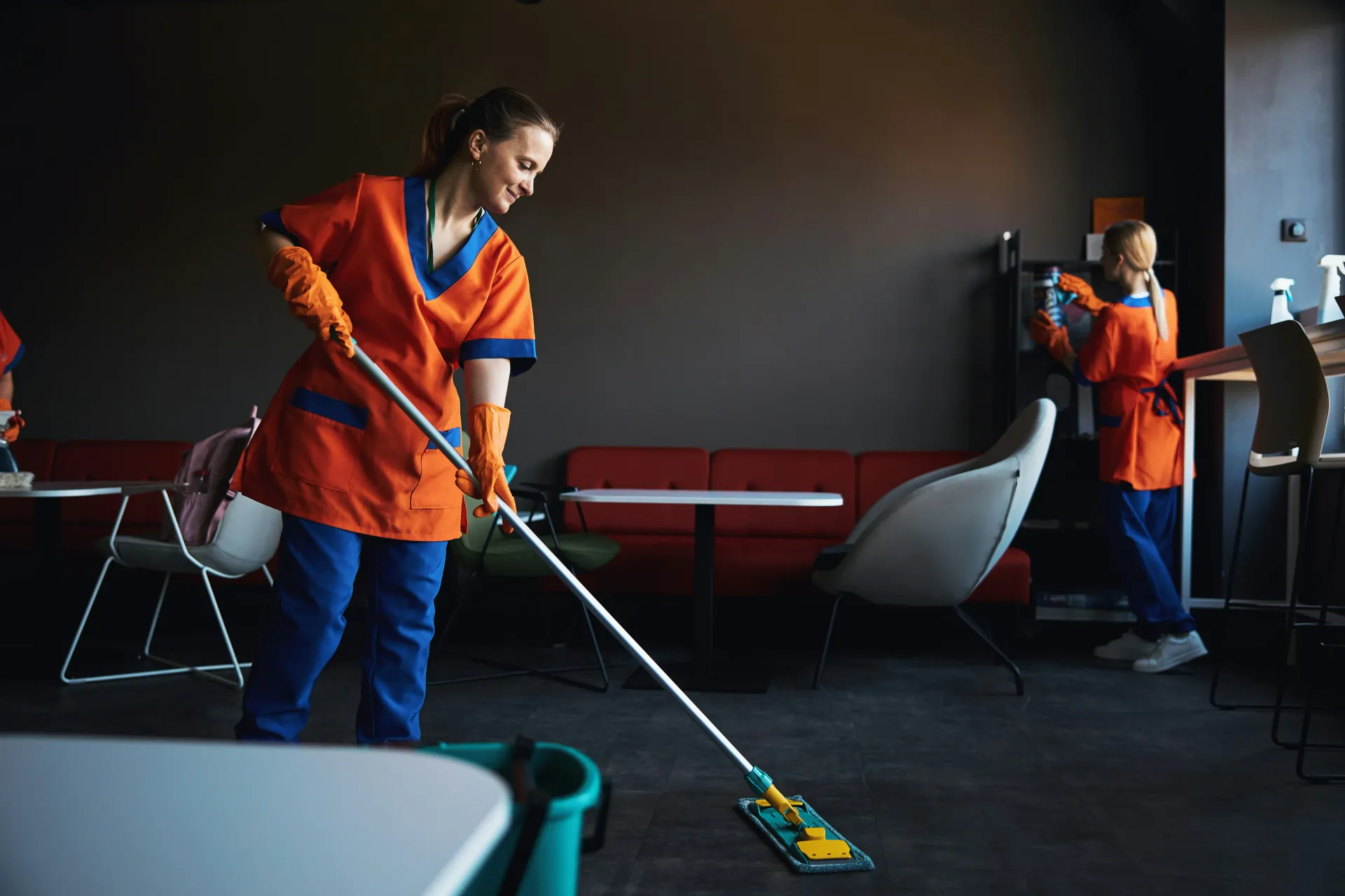 Two professional cleaners in uniform cleaning a modern office or commercial space, one mopping the floor and another wiping a surface.